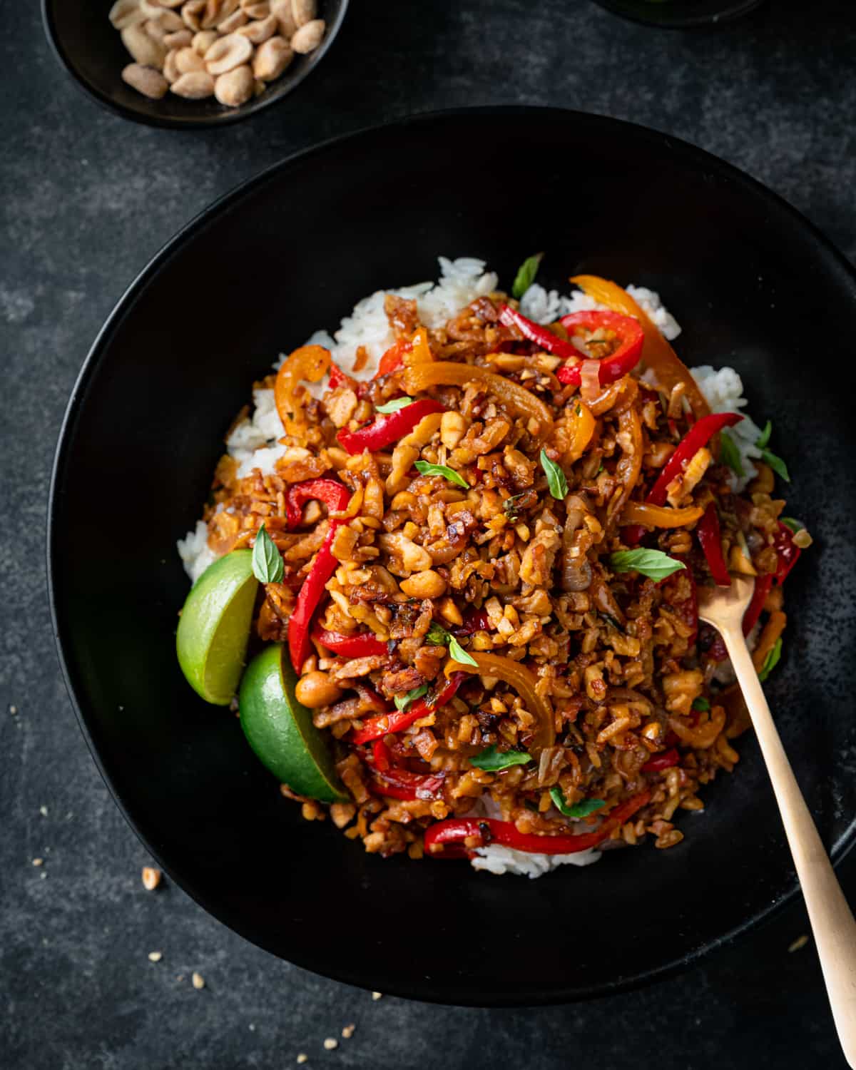 tempeh kecap stir fry in a black bowl over white rice with lime wedges.