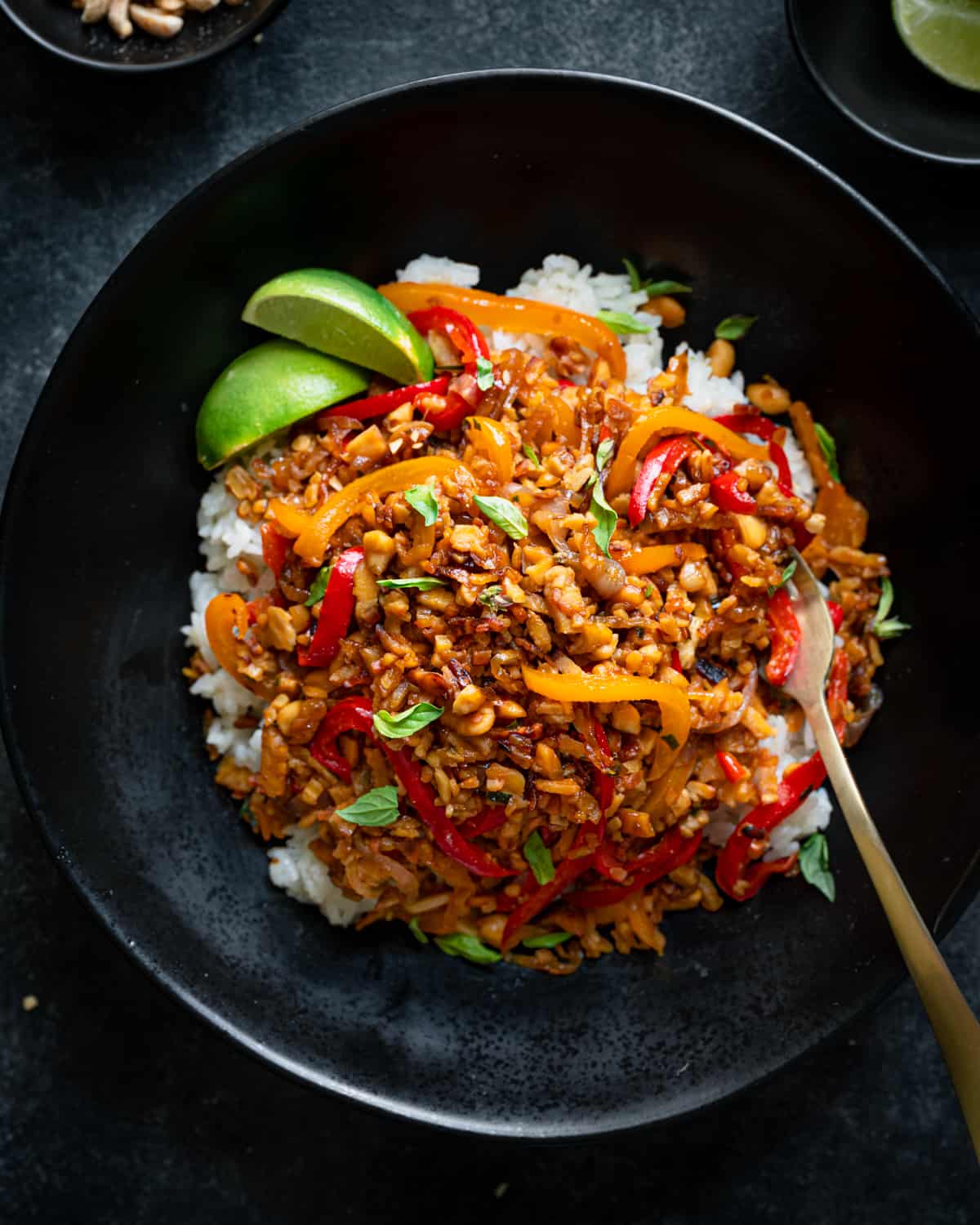 tempeh kecap stir fry in a black bowl over white rice with lime wedges and gold fork.