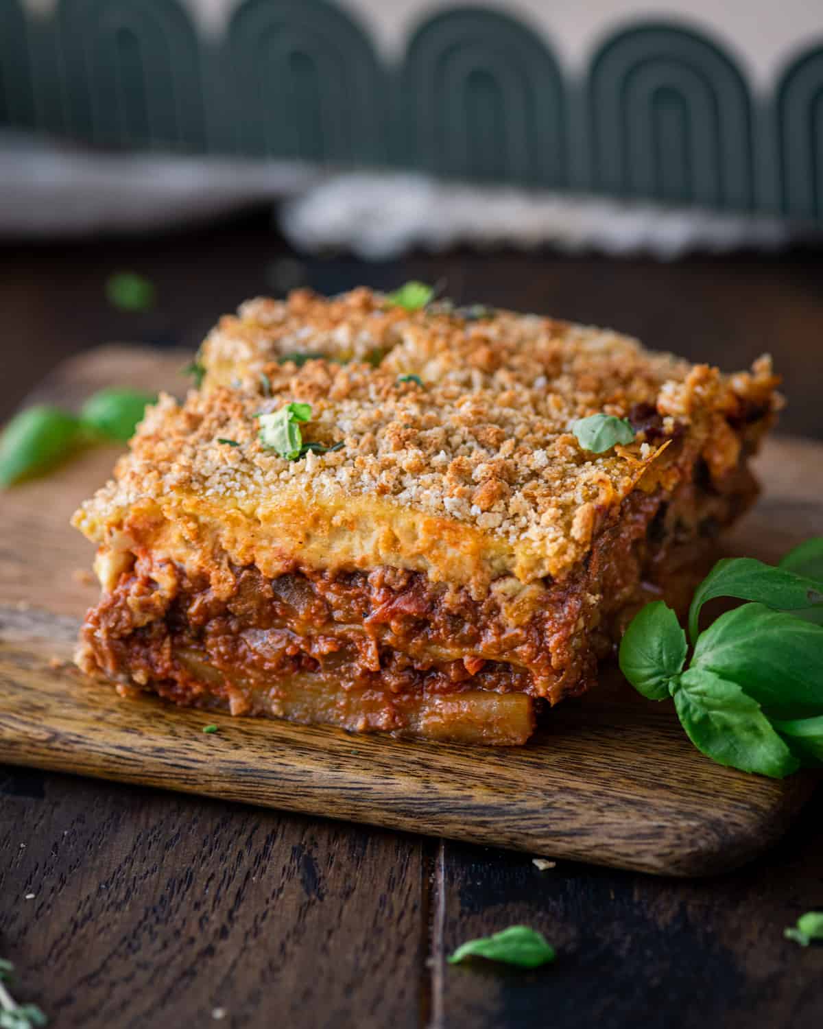 slice of vegan moussaka on a small wooden cutting board with fresh basil on the side.