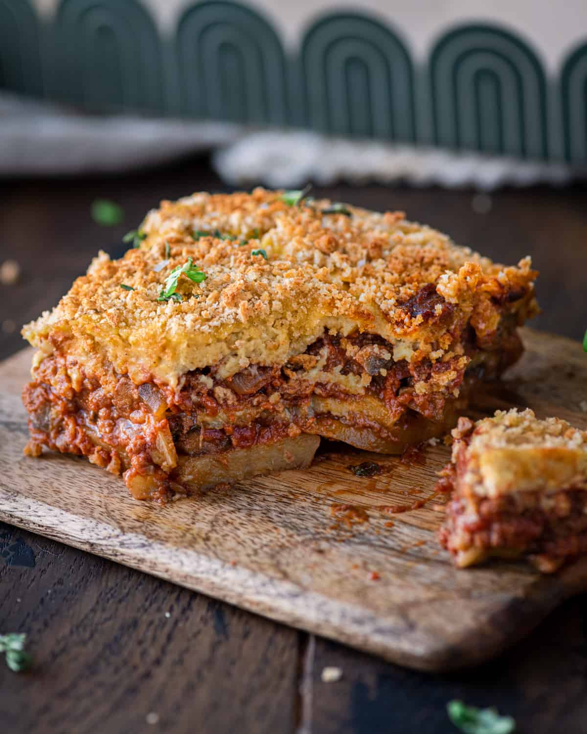 a big piece of vegan moussaka sliced so you can see the layers, sitting on a wooden cutting board.
