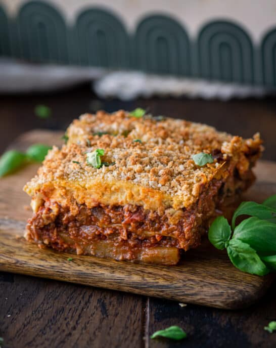 slice of vegan moussaka on a small wooden cutting board with fresh basil on the side.