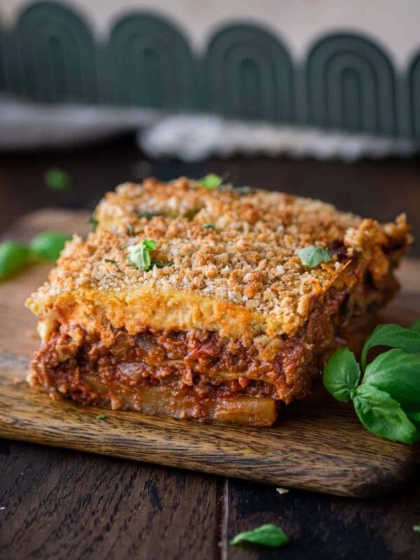 slice of vegan moussaka on a small wooden cutting board with fresh basil on the side.