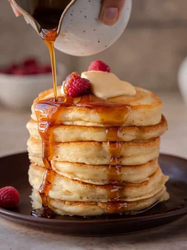 stack of fluffy vegan pancakes on a brown plate with maple syrup being drizzled on top.