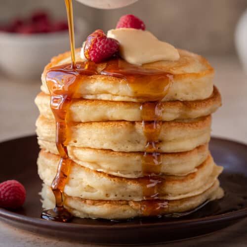 stack of fluffy vegan pancakes on a brown plate with maple syrup being drizzled on top.