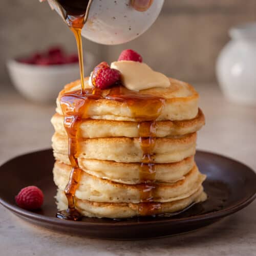 stack of fluffy vegan pancakes on a brown plate with maple syrup being drizzled on top.