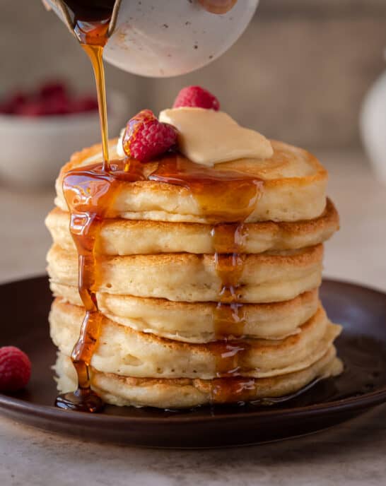 stack of fluffy vegan pancakes on a brown plate with maple syrup being drizzled on top.
