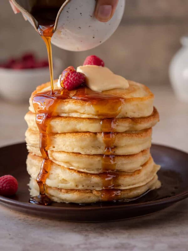 stack of fluffy vegan pancakes on a brown plate with maple syrup being drizzled on top.