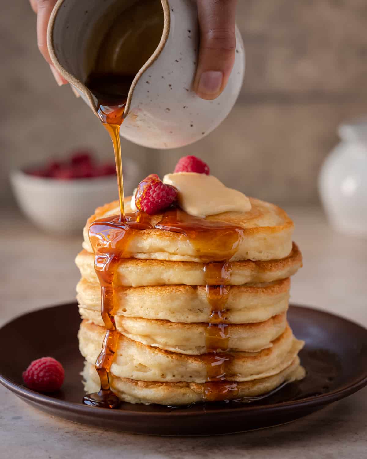 woman's hands pouring maple syrup onto a stack of fluffy golden vegan pancakes on a brown plate.