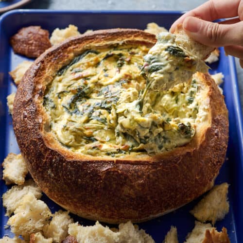 Woman dipping bread into spinach artichoke dip in a bread bowl.