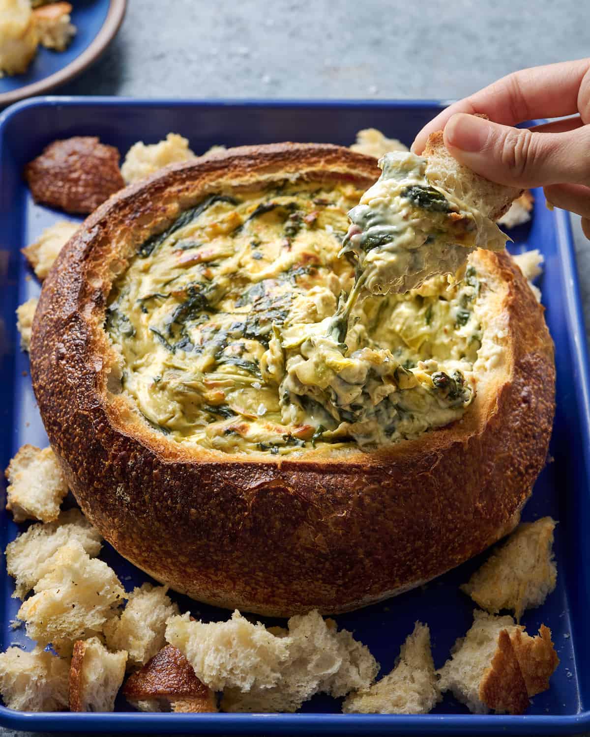 Woman dipping bread into spinach artichoke dip in a bread bowl.