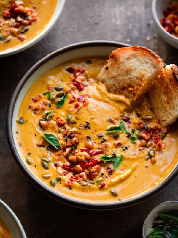 backlit angle of a bowl of thai pumpkin soup with a piece of toasted bread dunking in to show its creamy texture.