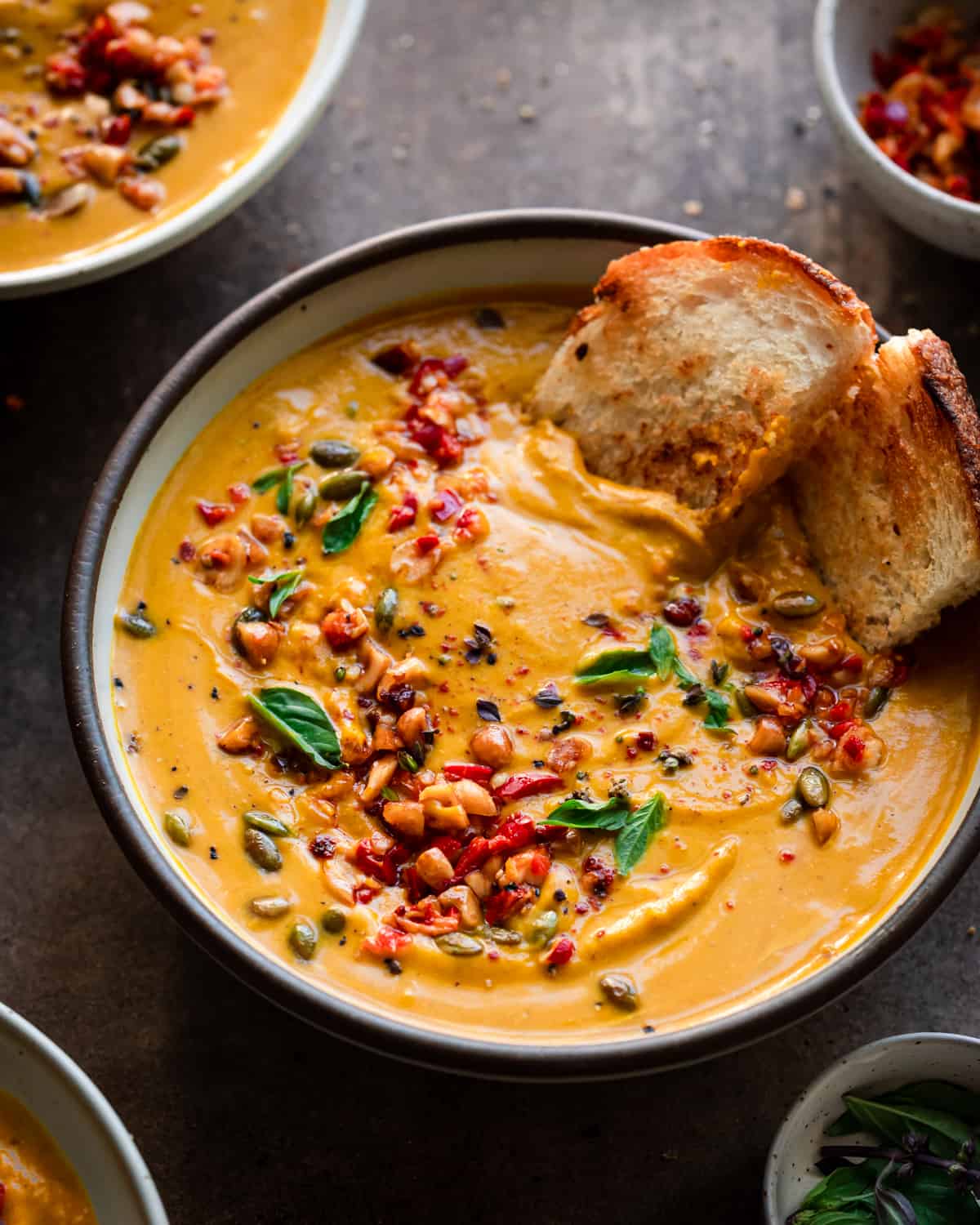 backlit angle of a bowl of thai pumpkin soup with a piece of toasted bread dunking in to show its creamy texture.