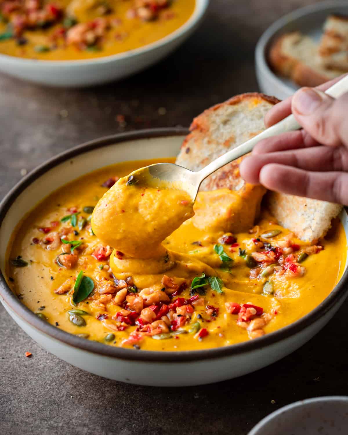 woman's hands spooning a creamy thai pumpkin soup from a ceramic bowl on a brown table.