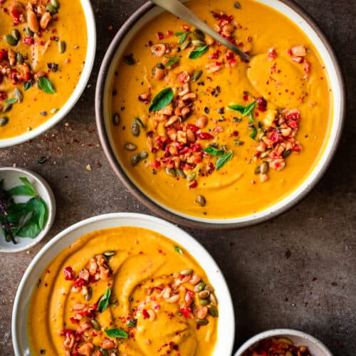 three bowls of thai pumpkin soup garnished with chiles and thai basil on a brown table.