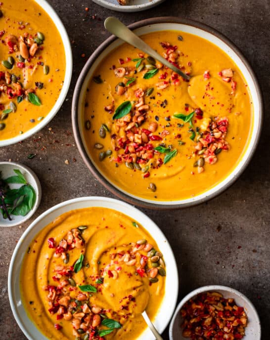 three bowls of thai pumpkin soup garnished with chiles and thai basil on a brown table.