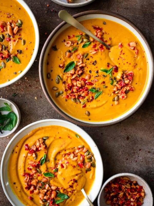 three bowls of thai pumpkin soup garnished with chiles and thai basil on a brown table.