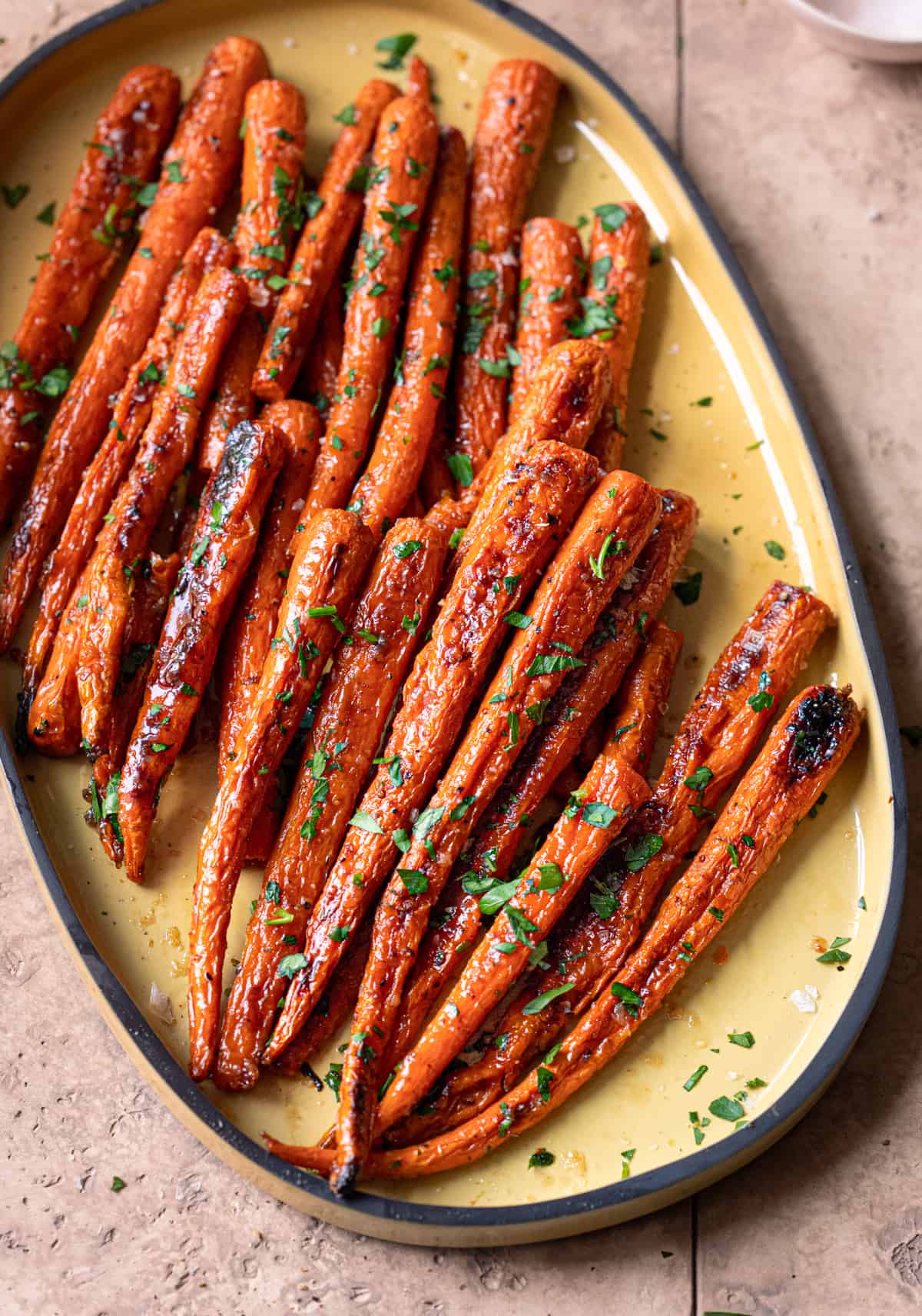 maple roasted carrots garnished with parsley on a yellow serving tray on a light pink tiled surface.