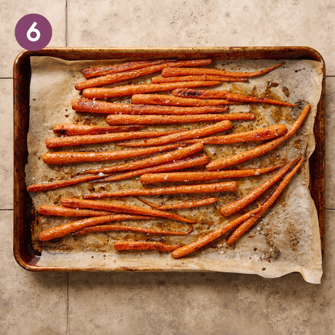 maple glazed carrots sitting on a parchment paper lined sheet pan on a beige tiled surface. 