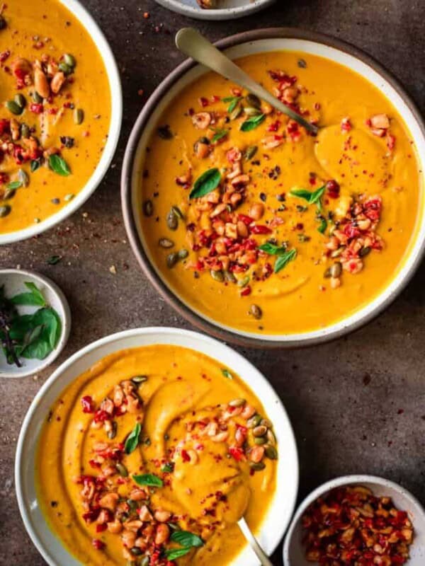 three bowls of thai pumpkin soup garnished with chiles and thai basil on a brown table.