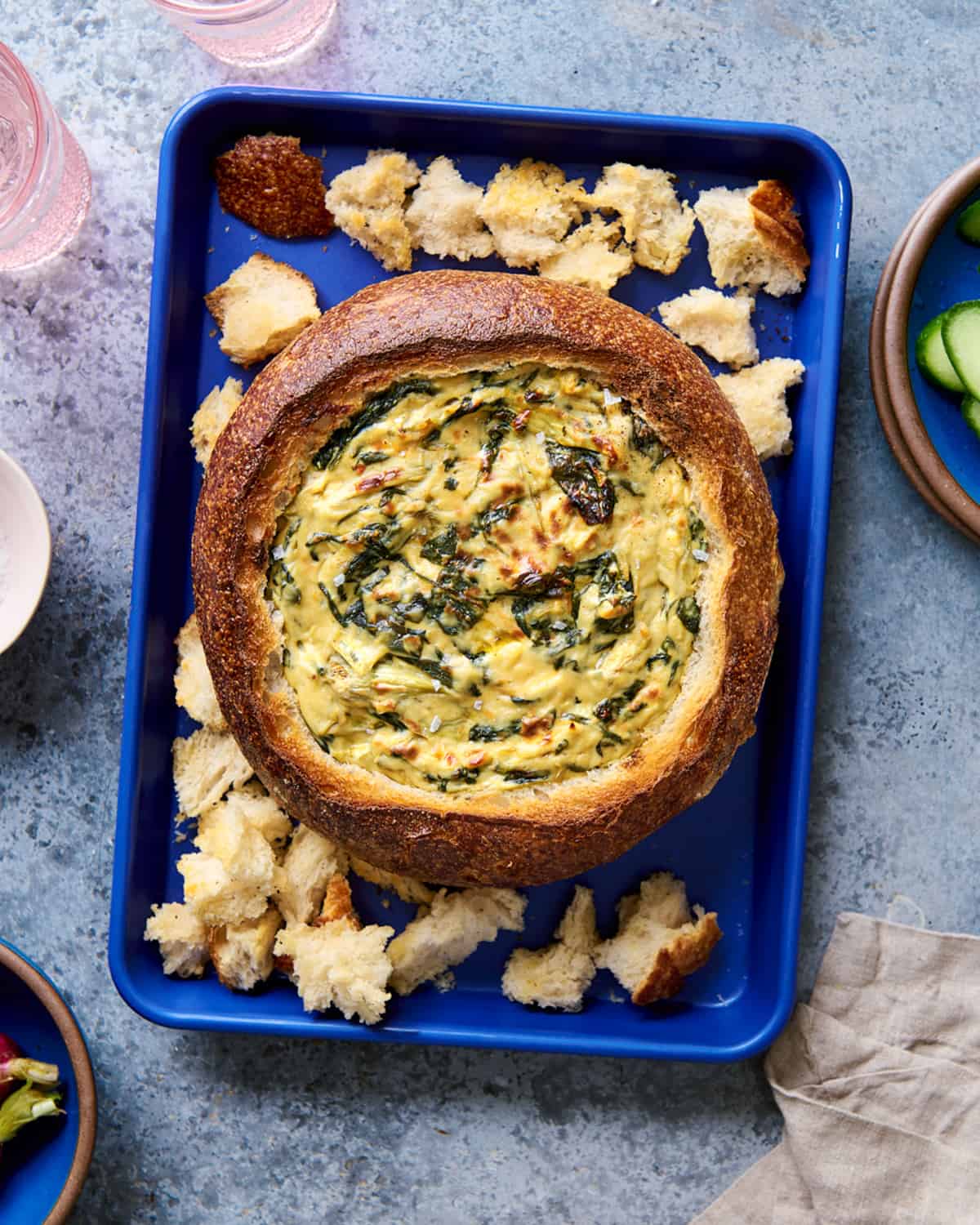 vegan spinach artichoke dip in bread bowl next to cooked bread pieces on a blue sheet pan.