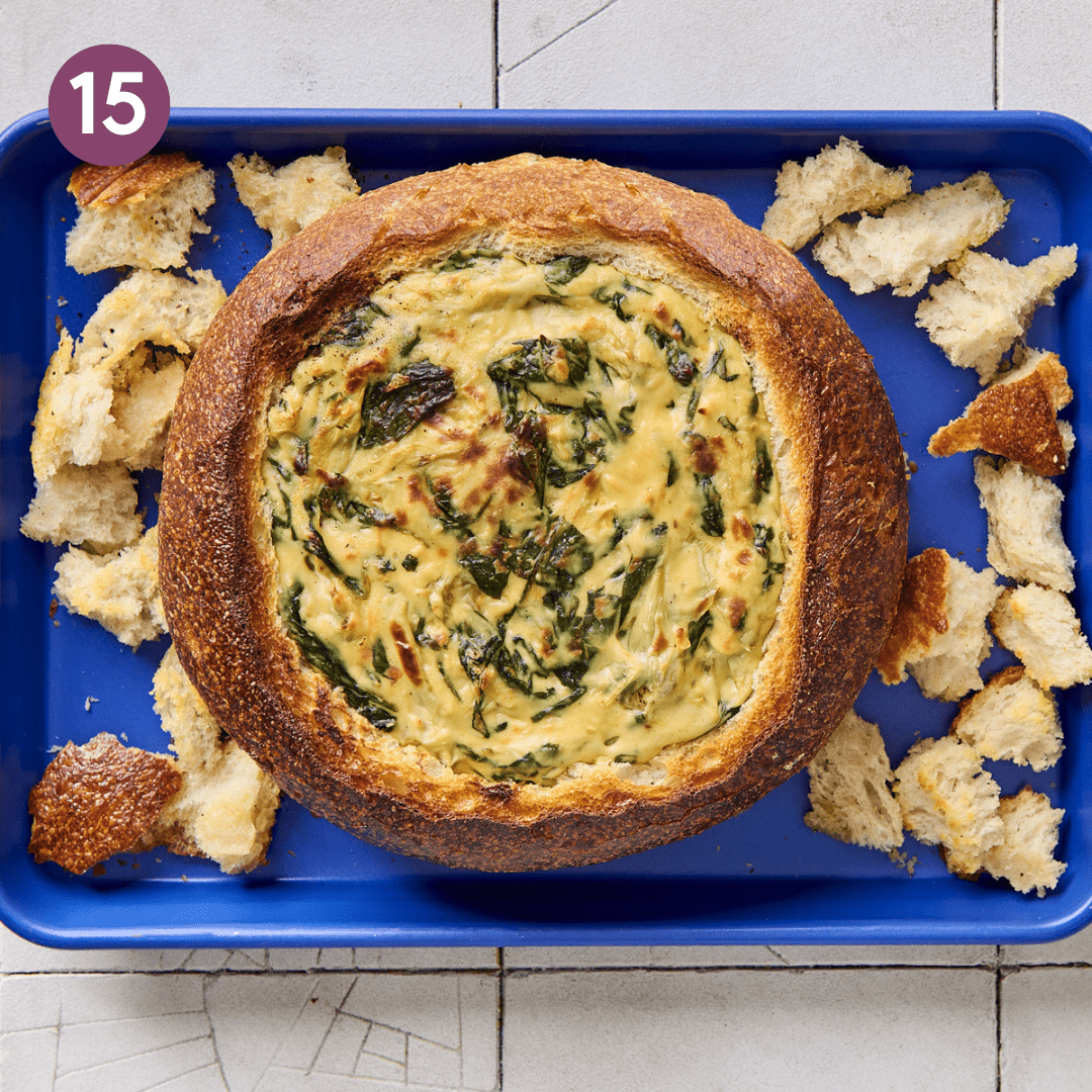 Bread bowl filled with spinach artichoke dip and croutons on sheet pan after being cooked.