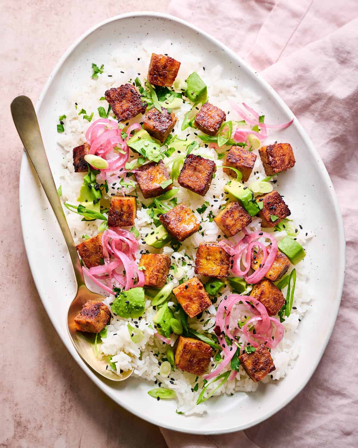 marinated tofu cubes on a platter of white rice with scallions and pickled onions on top of a pink linen.