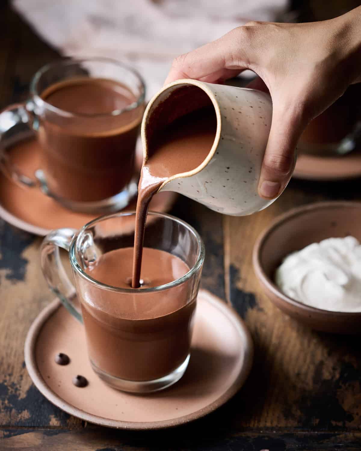Person pouring hot chocolate into glass mug on wood table.