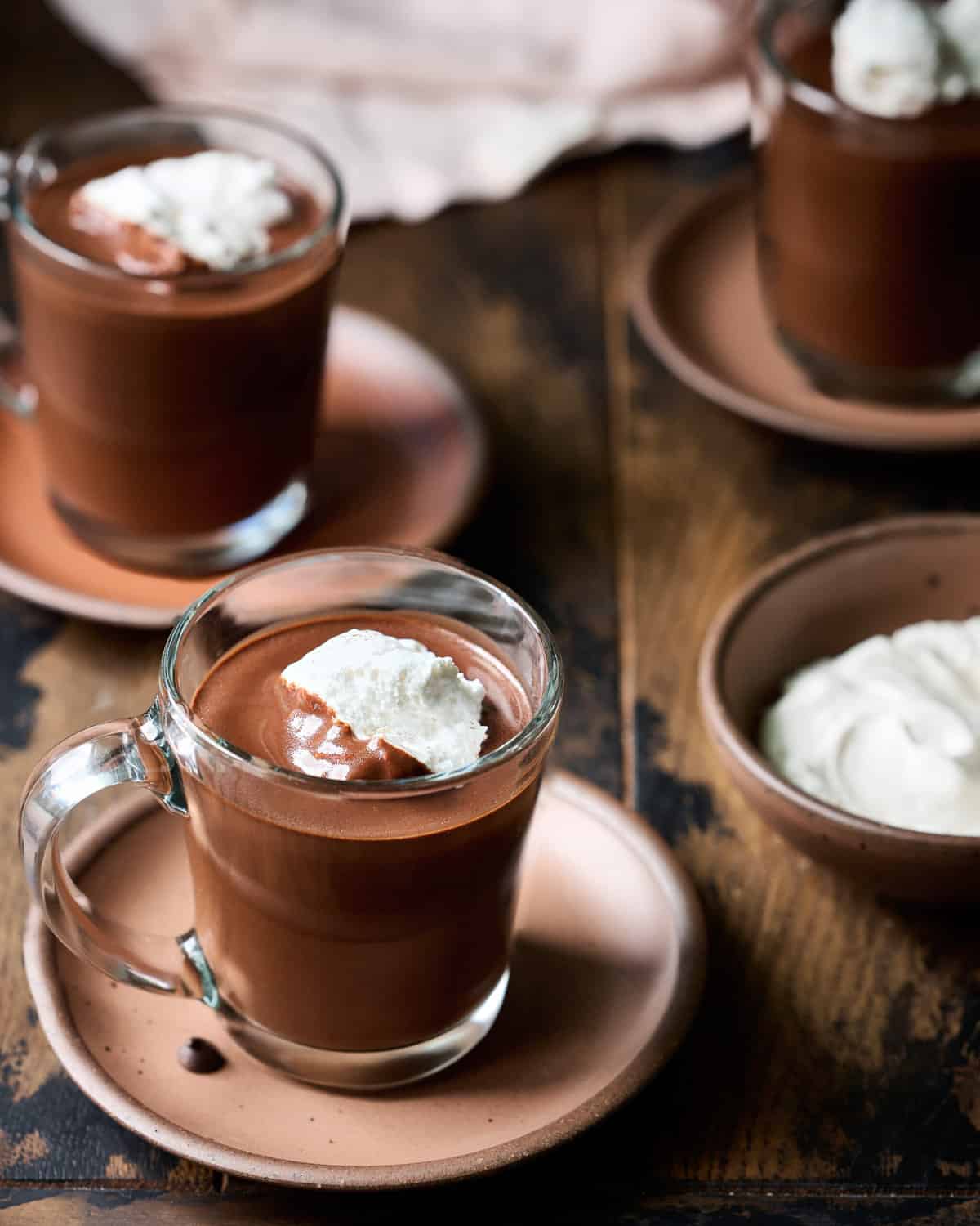 Three glass mugs filled with hot chocolate and whipped cream on plates on a wood table.