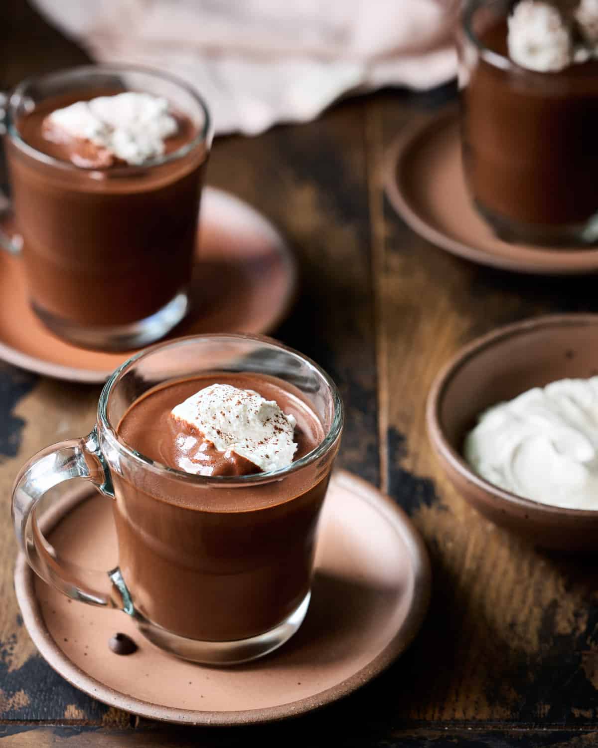 Three glass mugs filled with hot chocolate and whipped cream on plates on a wood table.