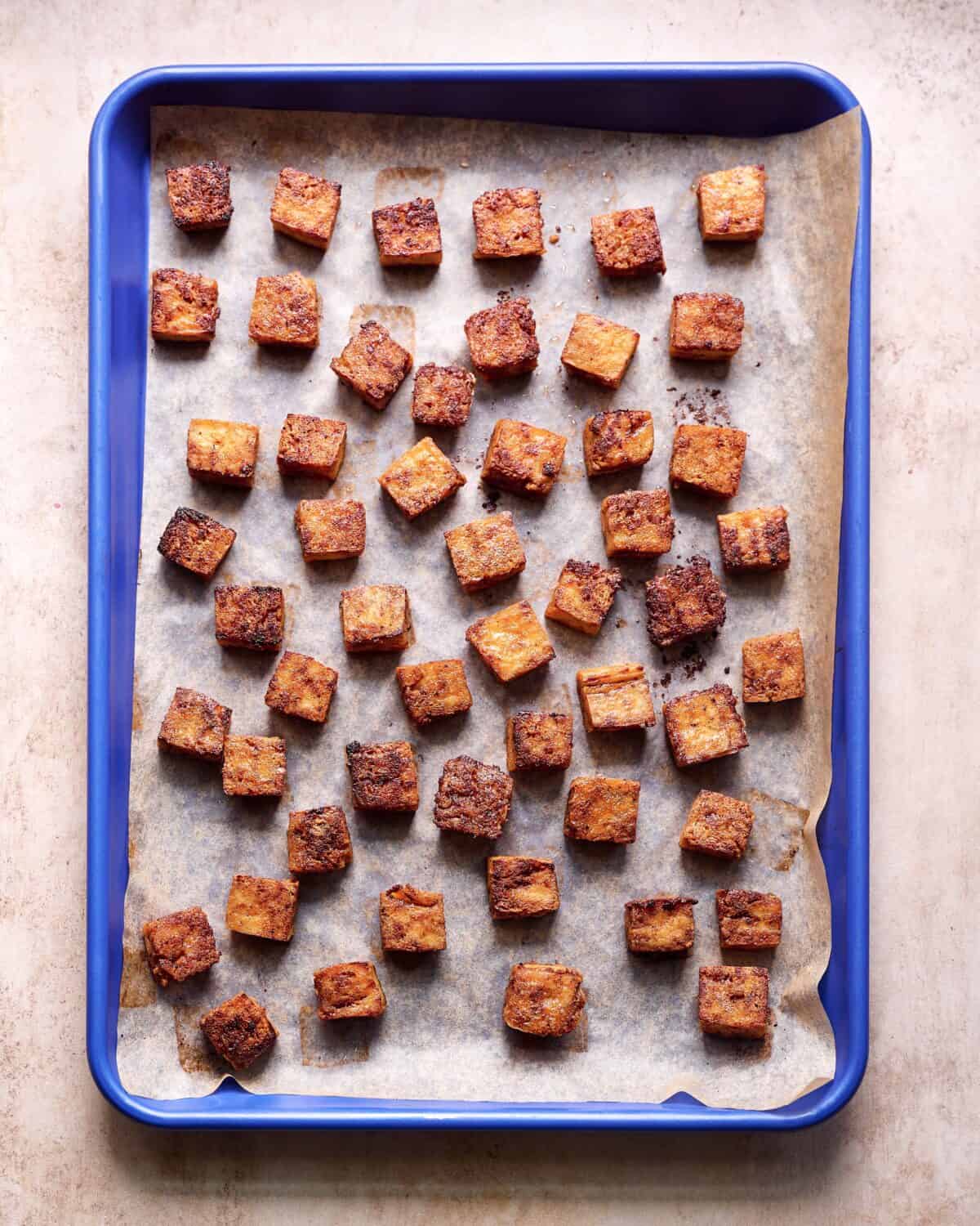 Crispy marinated tofu cubes on a parchment paper lined blue sheet pan on a pink surface.