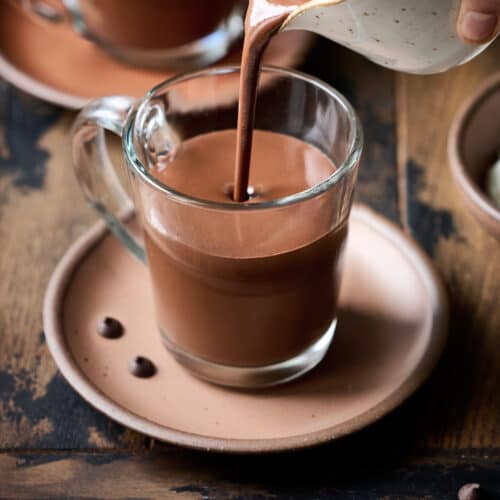 Person pouring hot chocolate into a glass mug on a wood table.