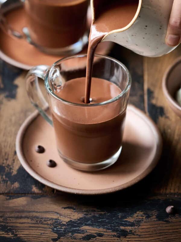 Person pouring hot chocolate into a glass mug on a wood table.