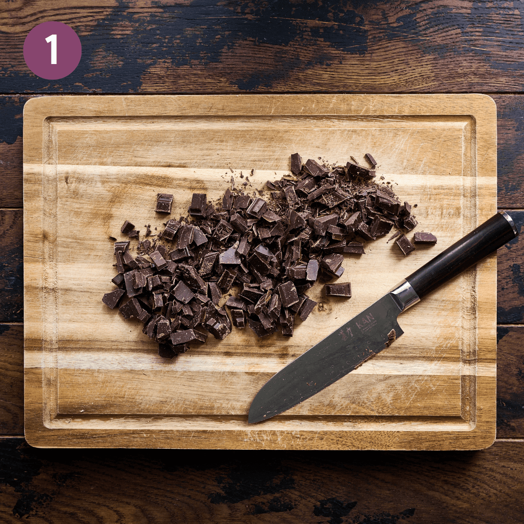 Knife with chopped dark chocolate on a wood cutting board.