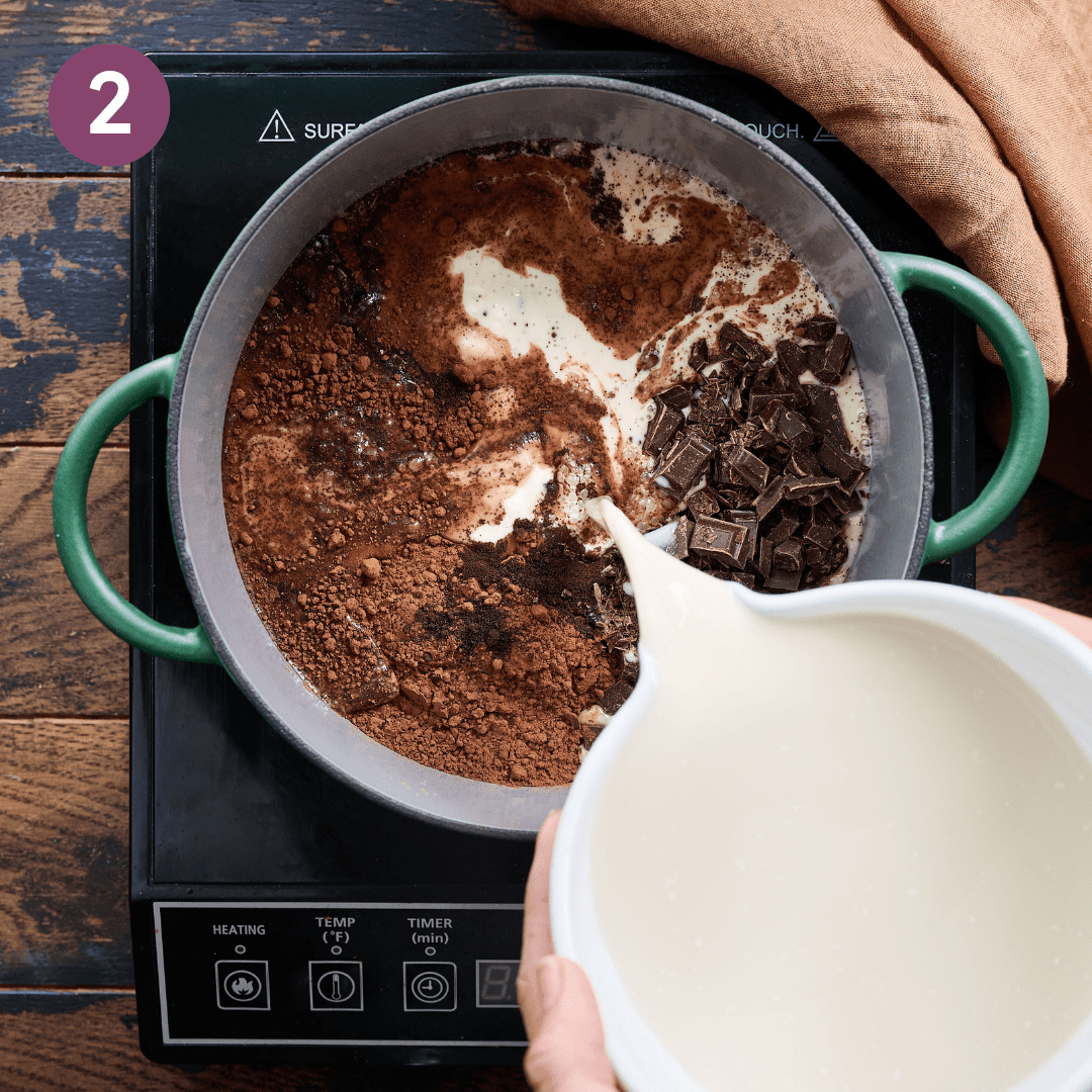 Oat and coconut milk being poured into the rest of hot chocolate ingredients in a pot on a portable stovetop.