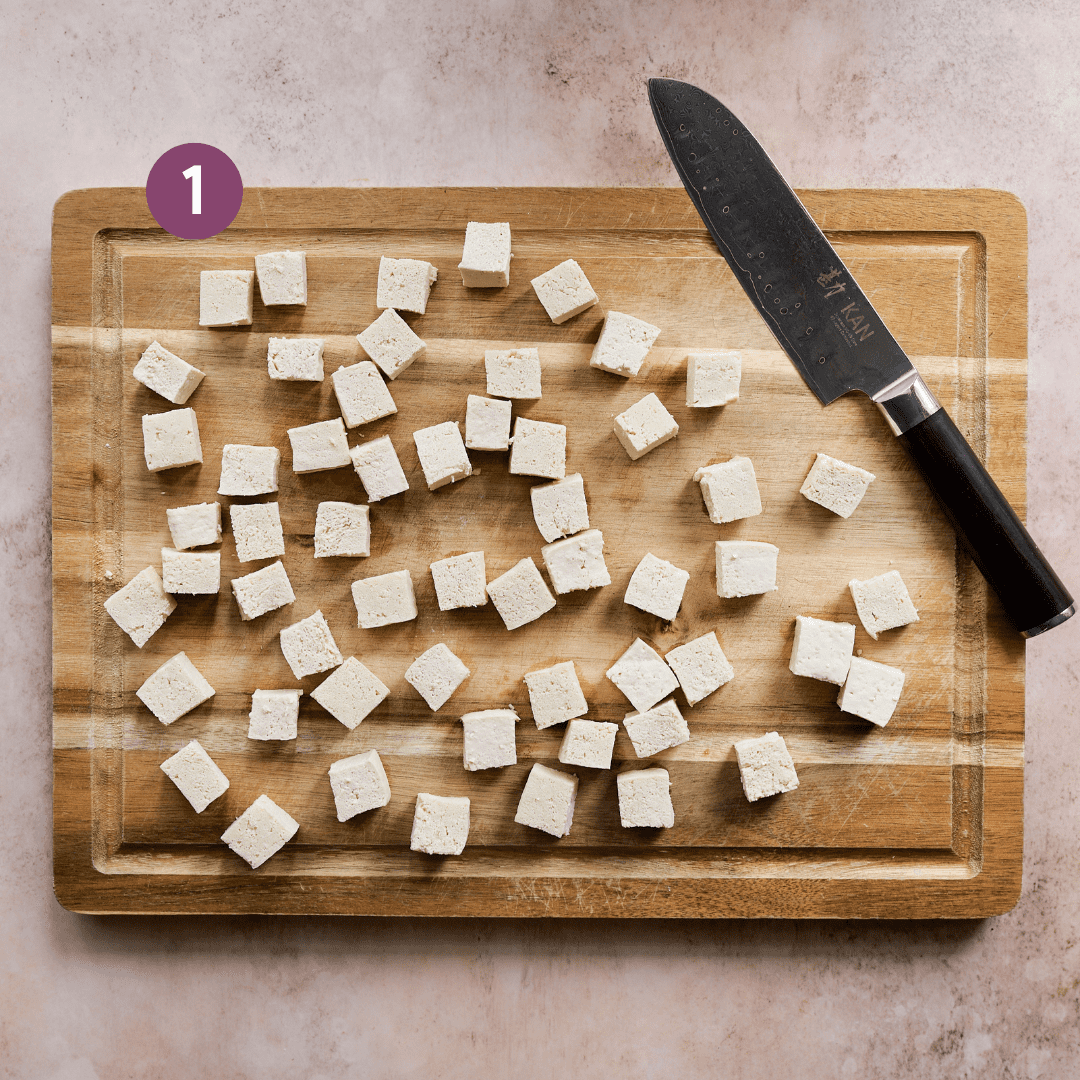 sliced tofu cubes on a wooden cutting board with a knife on a pink table.