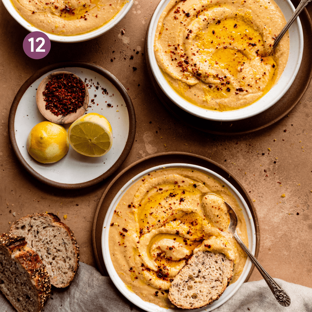 3 bowls of creamy vegan cauliflower soup on a brown table with linens, lemons, chile flakes, and bread on the side.