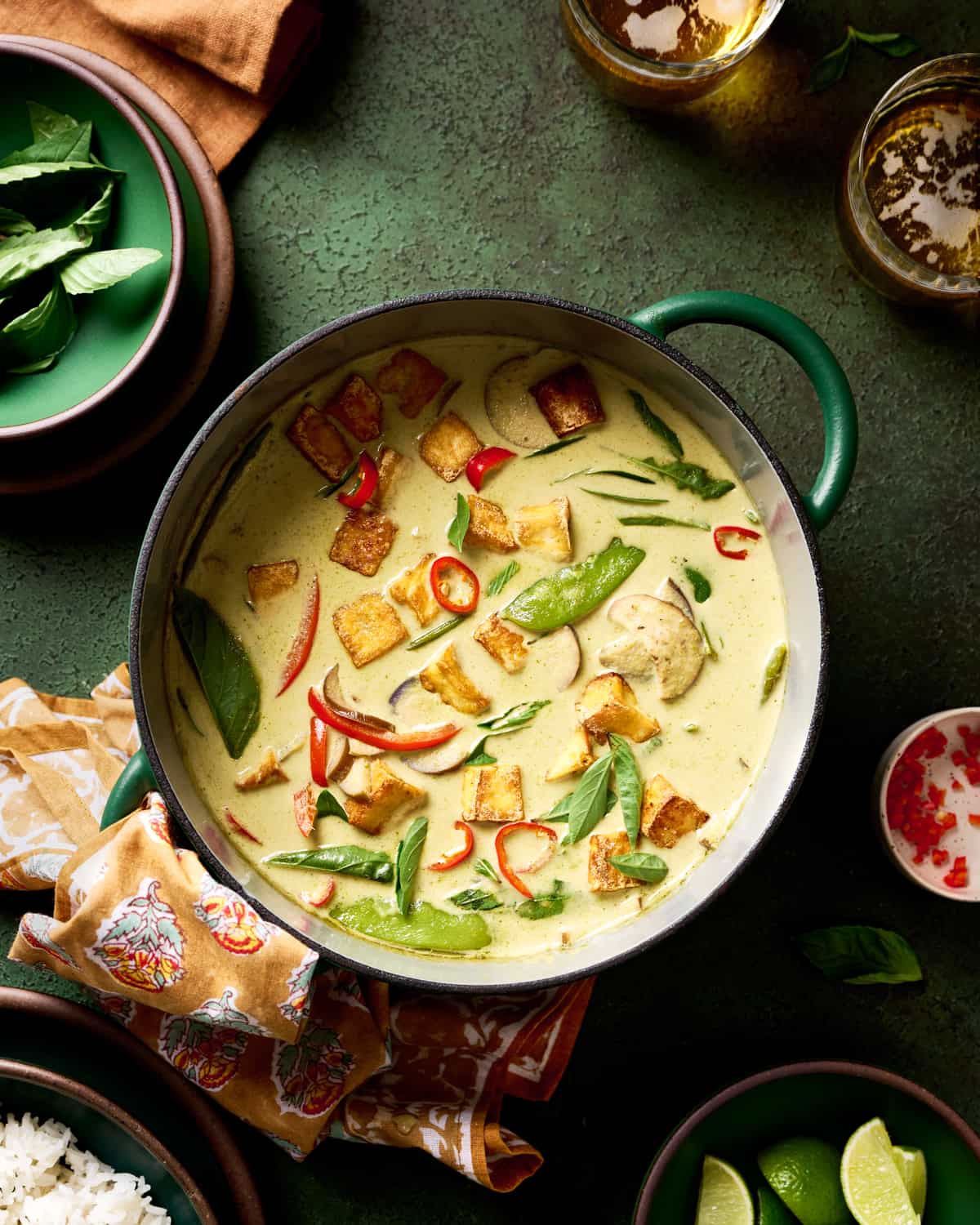 thai vegan green curry with crispy tofu and vegetables in a green saucepan on a green table with beer and limes for serving.