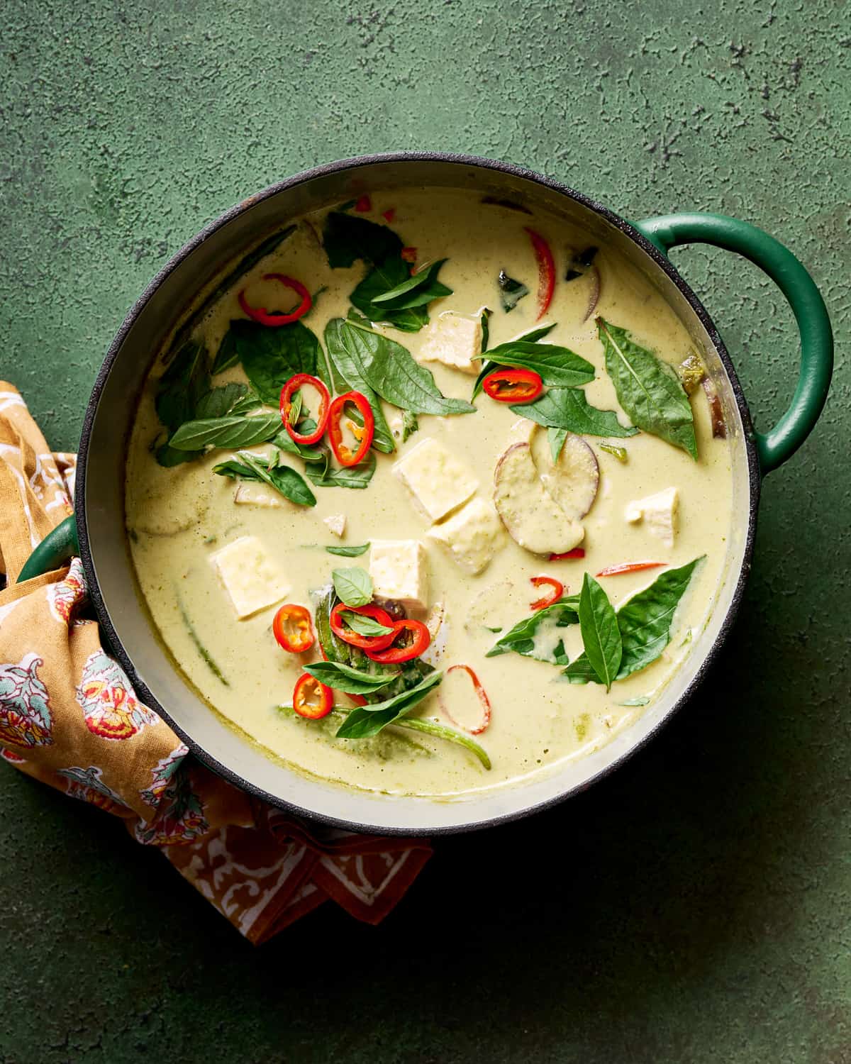 Thai green curry with tofu and vegetables and thai basil in a green saucepan on a green table.