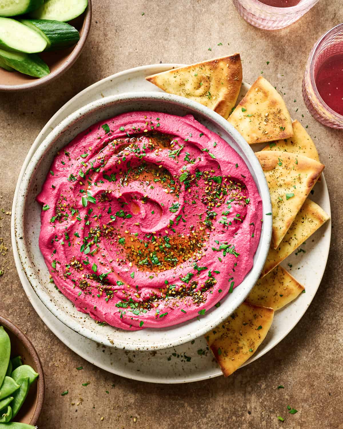 Bowl of beet hummus on a plate with pita bread next to bowls of cucumbers and snap peas and two glasses of juice.