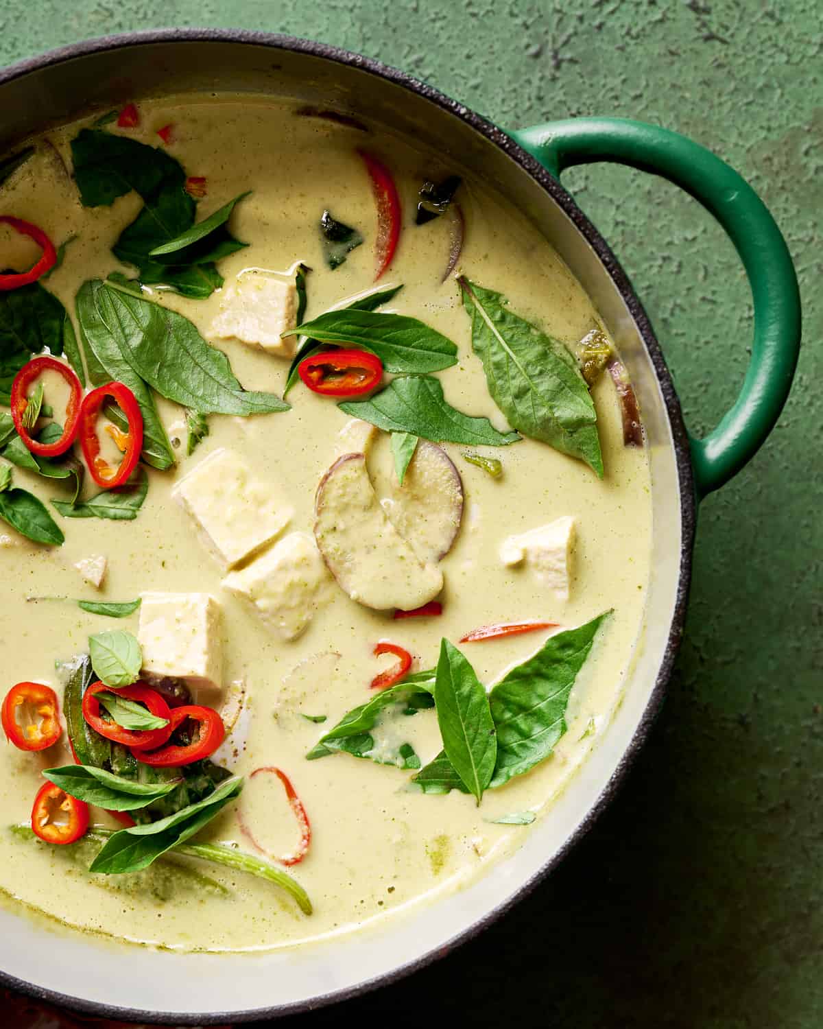 closeup photo of Thai green curry with tofu and vegetables and thai basil in a green saucepan.