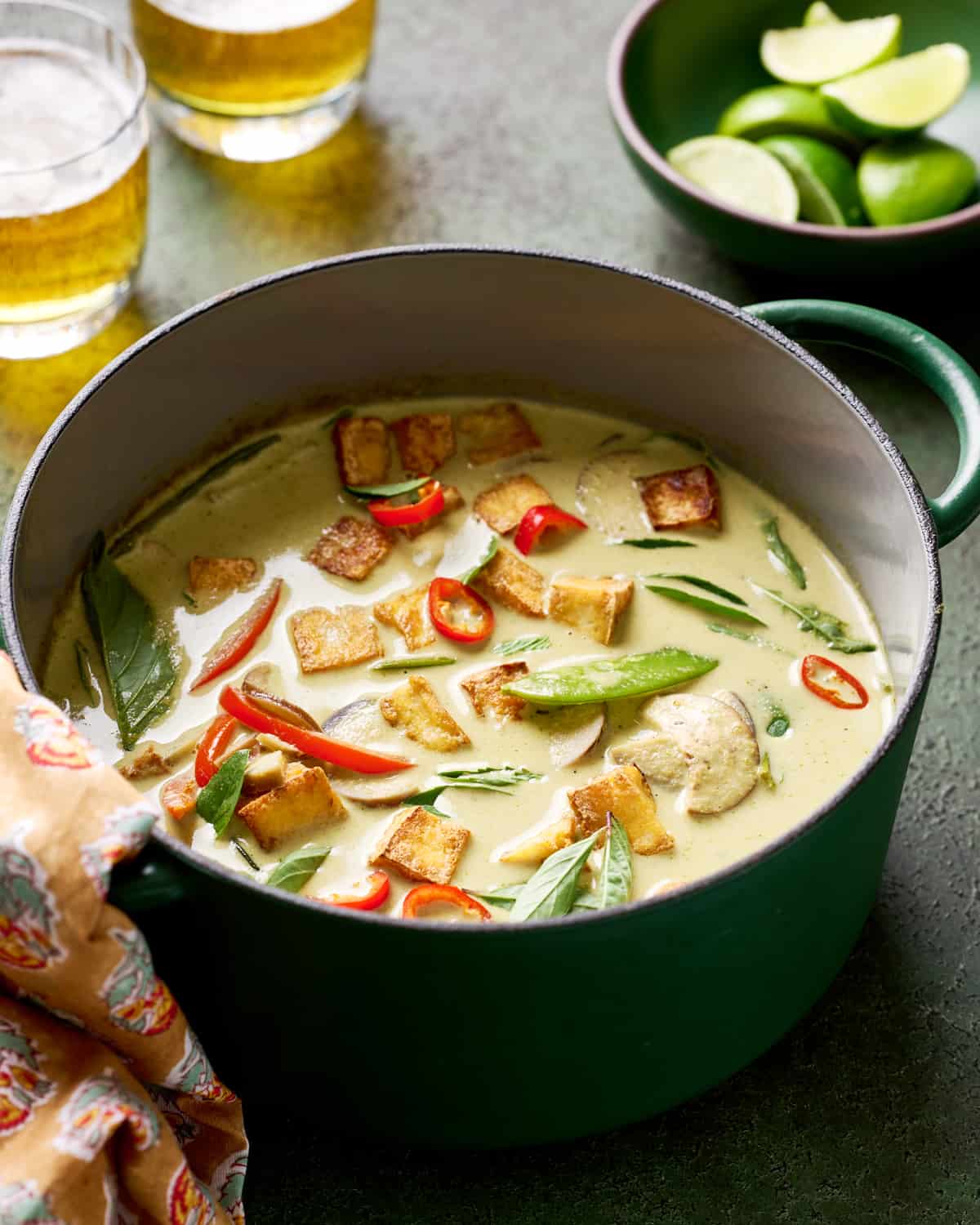 backlit shot of Thai vegan green curry with crispy tofu and vegetables in a green saucepan with beer and limes in background.