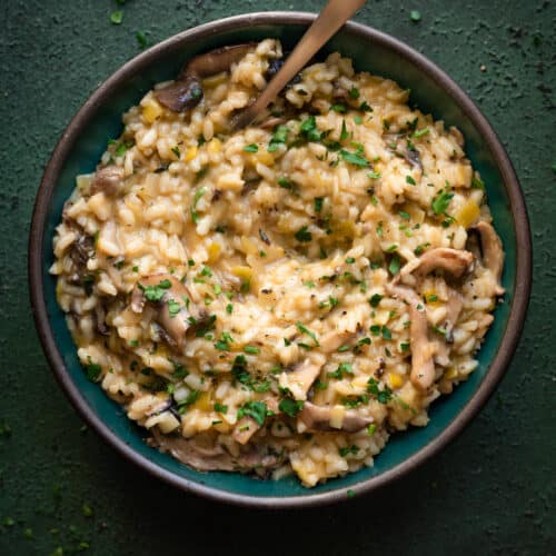 creamy, velvety vegan mushroom risotto in a green bowl with spoon dug in on a dark green table.
