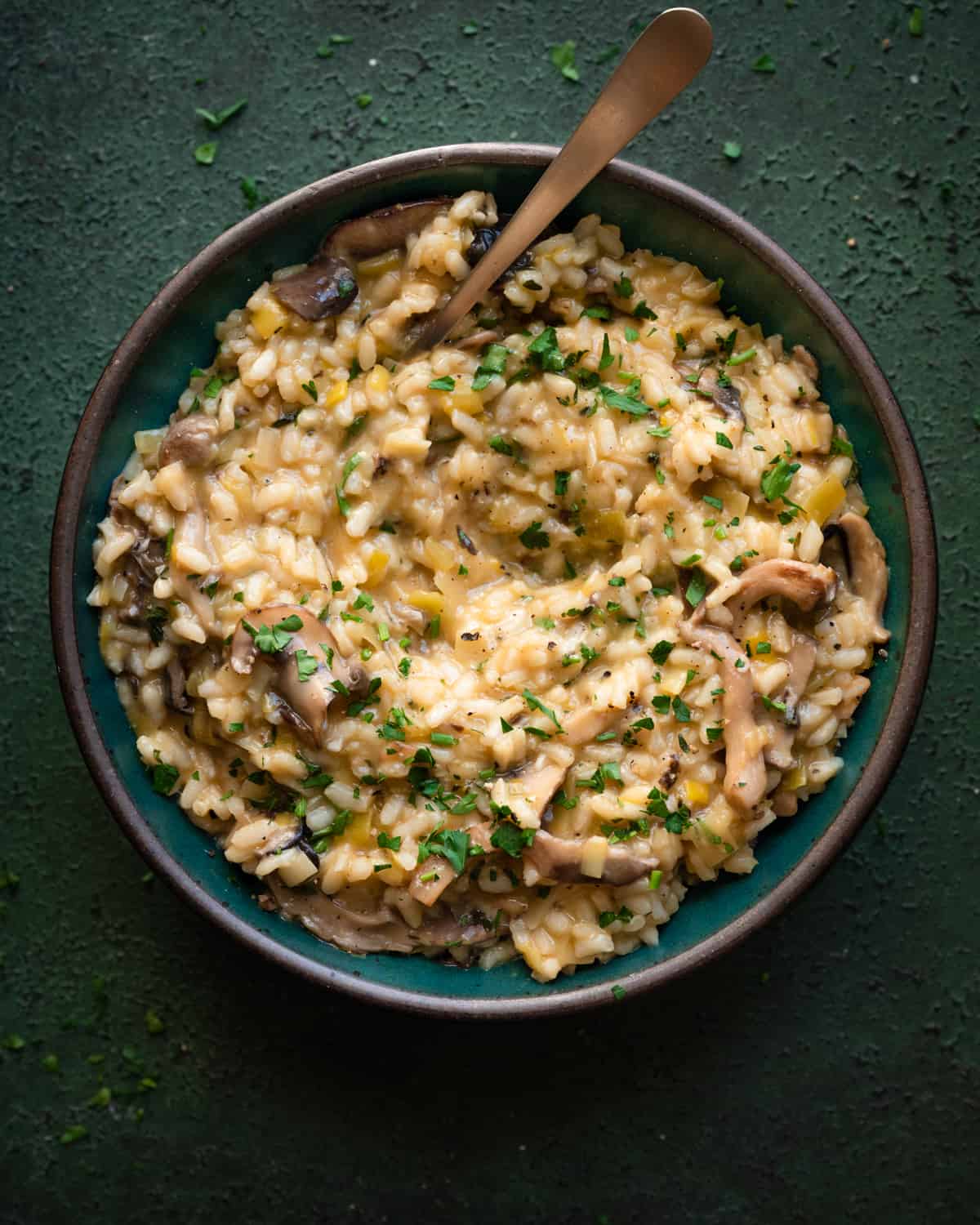 creamy, velvety vegan mushroom risotto in a green bowl with spoon dug in on a dark green table. 