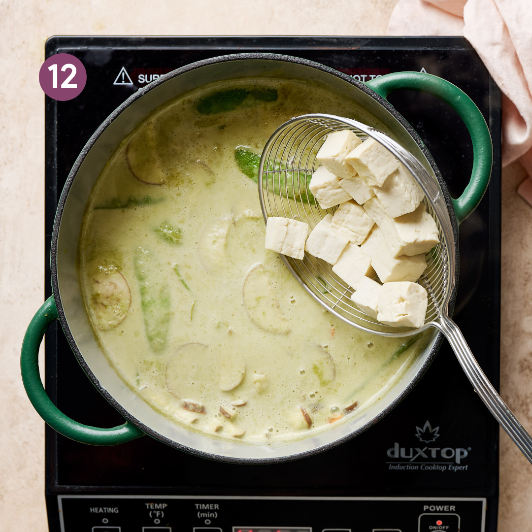slotted spoon adding tofu cubes to a vegan thai green curry in a green saucepan.