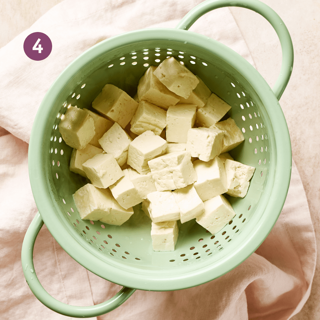 tofu cubes that have been boiled sitting in a light green colander on a light pink surface. 