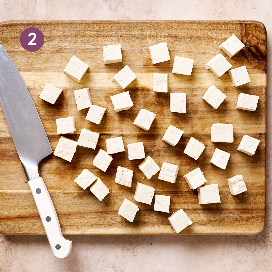 tofu cubes sitting on a wooden cutting board with a chef's knife on a light pink surface.