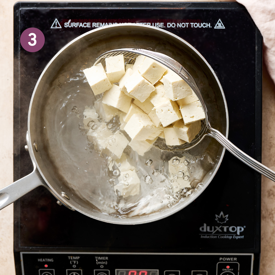 slotted spoon with tofu cubes being lowered into a saucepan of boiling water.