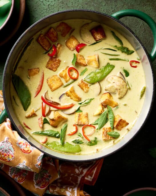 vegan thai green curry with vegetables and crispy tofu in a green saucepan on a green table.