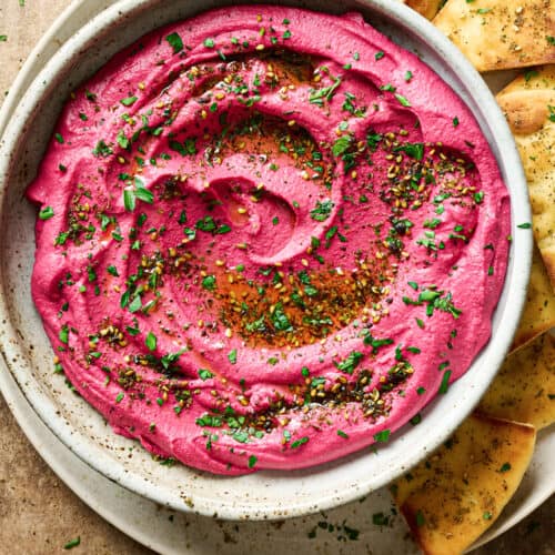 Vibrant pink beet hummus in a ceramic bowl, topped with parsley and za'atar on a plate with pita bread.