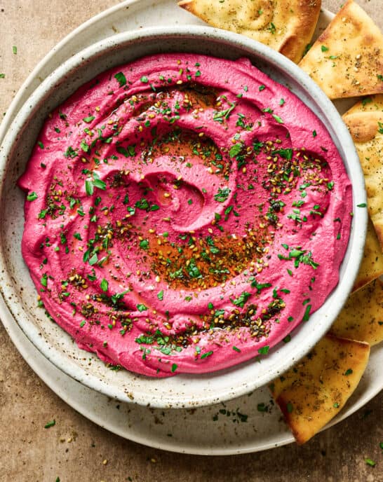 vibrant pink beet hummus in a ceramic bowl, topped with parsley and za'atar, on a plate with pita bread.
